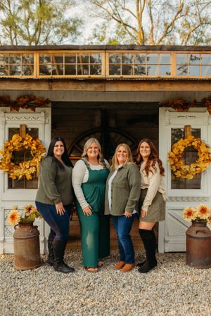 Four people pose in front of a rustic storefront decorated with fall wreaths and pumpkins.