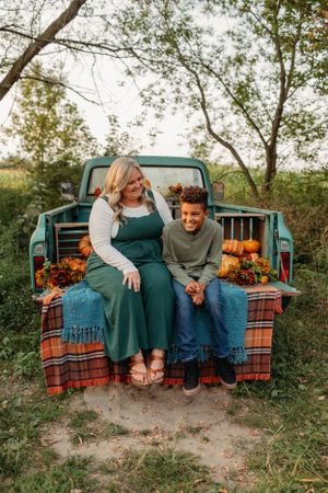 Two people sit on a vintage truck bed filled with pumpkins and plaid blankets.