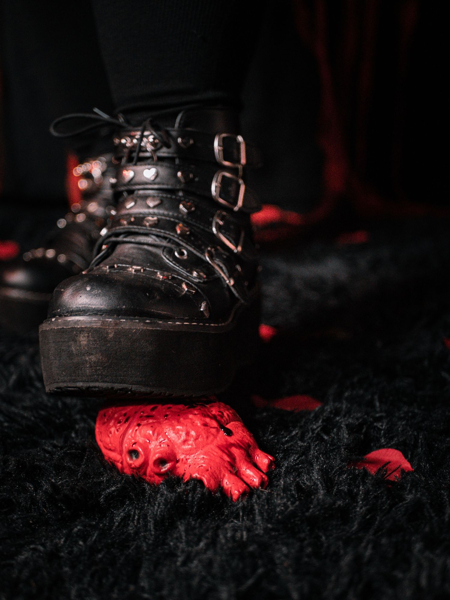 A dark leather combat boot with metal buckles sits next to a red plastic toy skeleton part against a black textured surface.