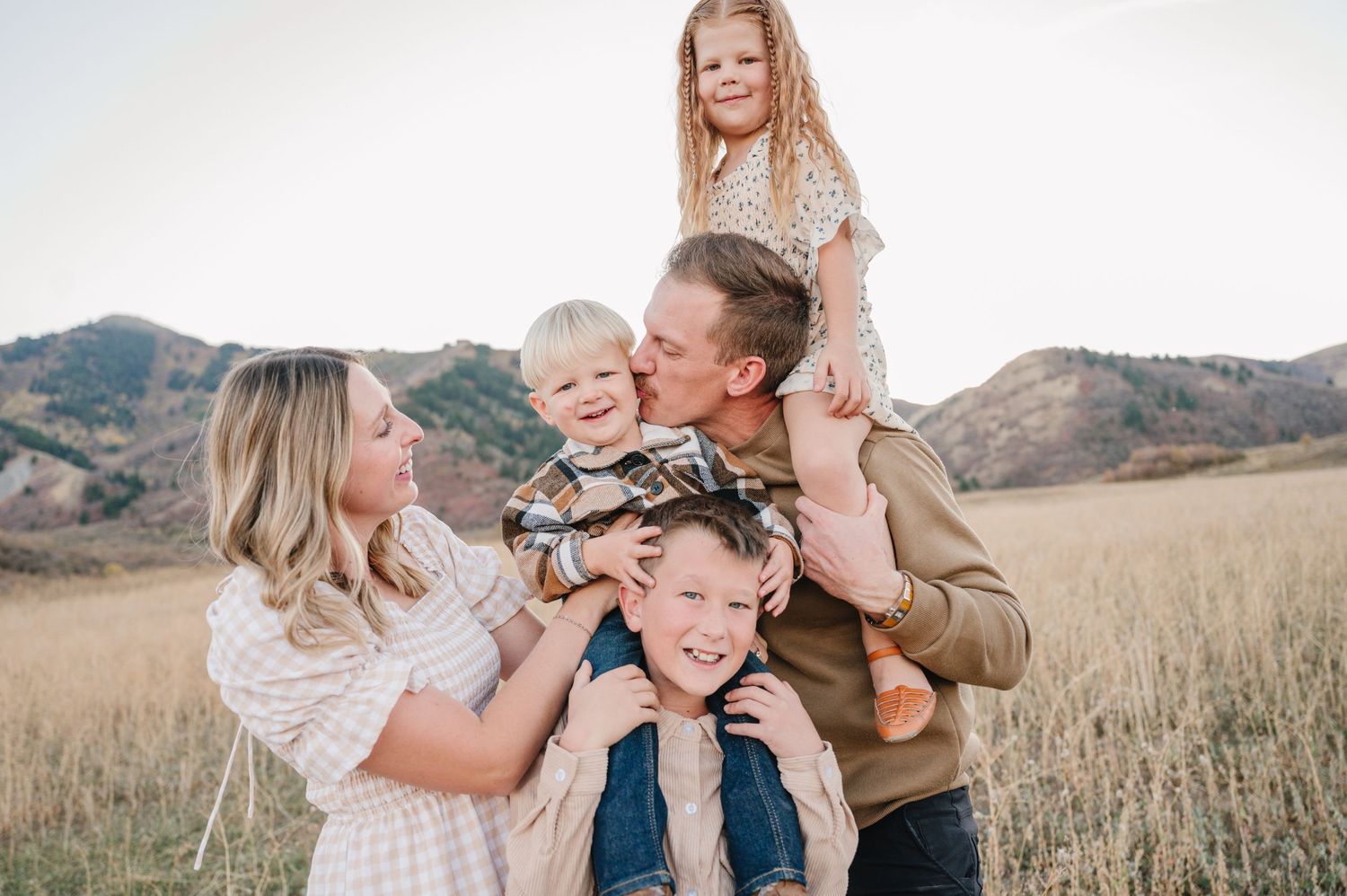A joyful family shares laughter and embraces while posing together in a golden field with mountains in the background.