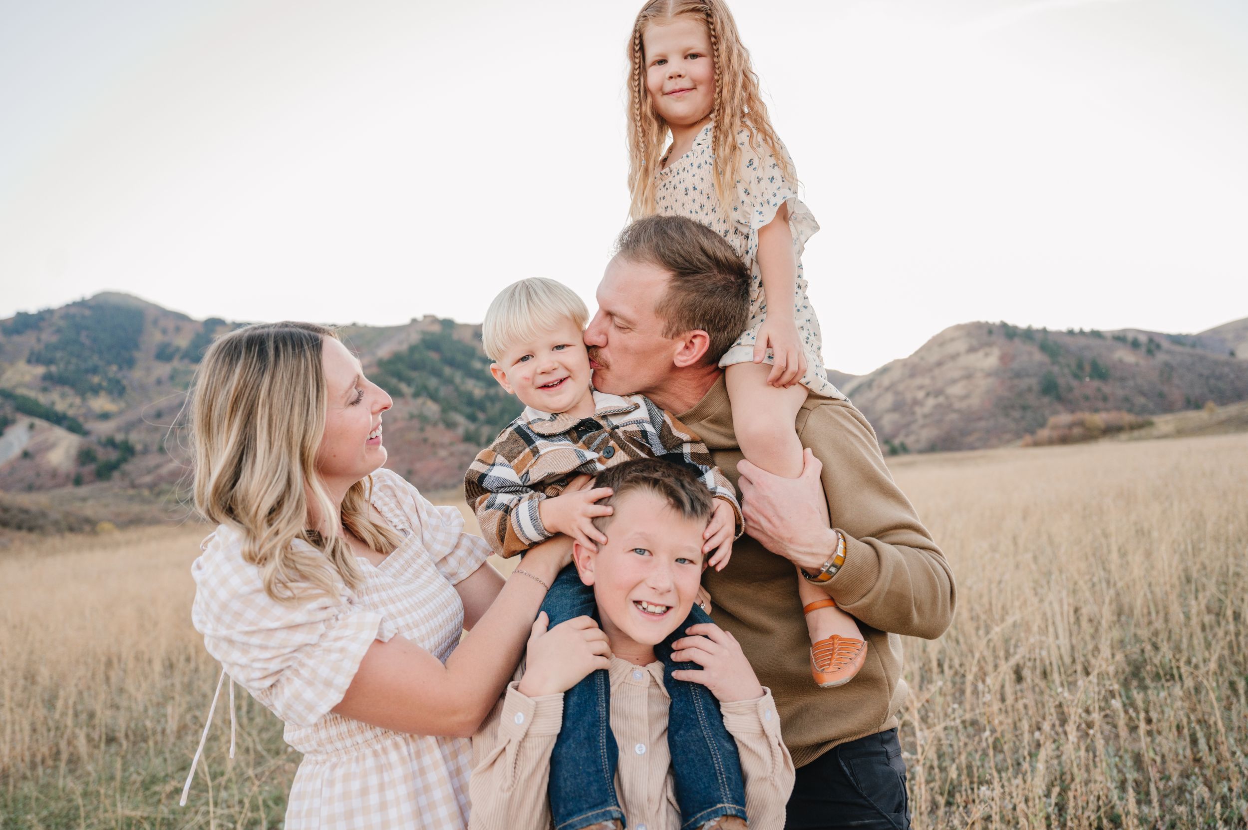 A joyful family shares laughter and embraces while posing together in a golden field with mountains in the background.