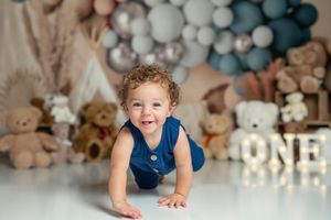 Baby in blue outfit crawls on white floor with balloon decorations and teddy bears in background.