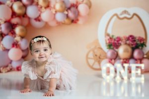 First birthday photo with pink balloon arch and illuminated ONE sign in princess theme setting.