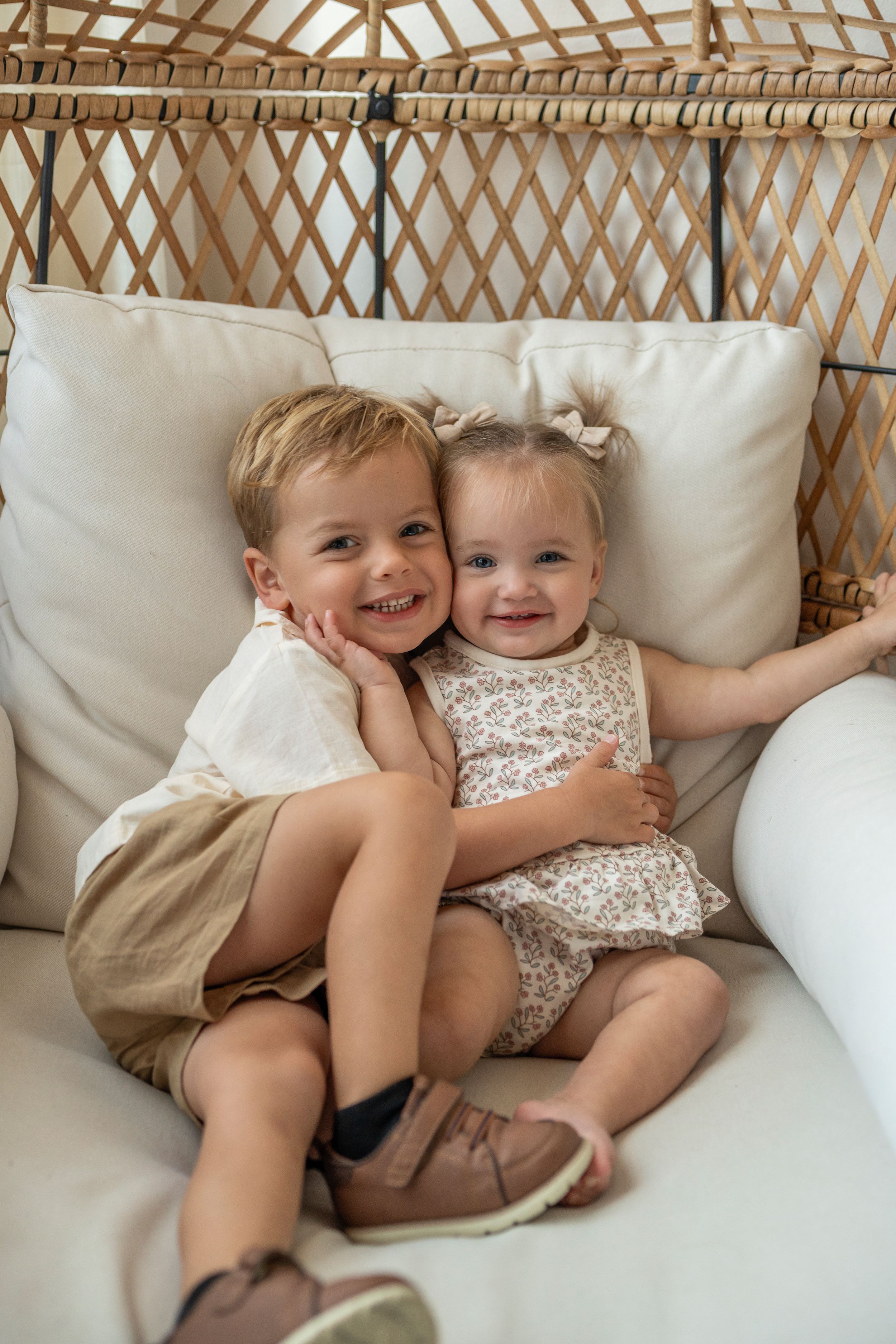 Two children share joyful moments while cuddling together on a white couch in matching outfits.