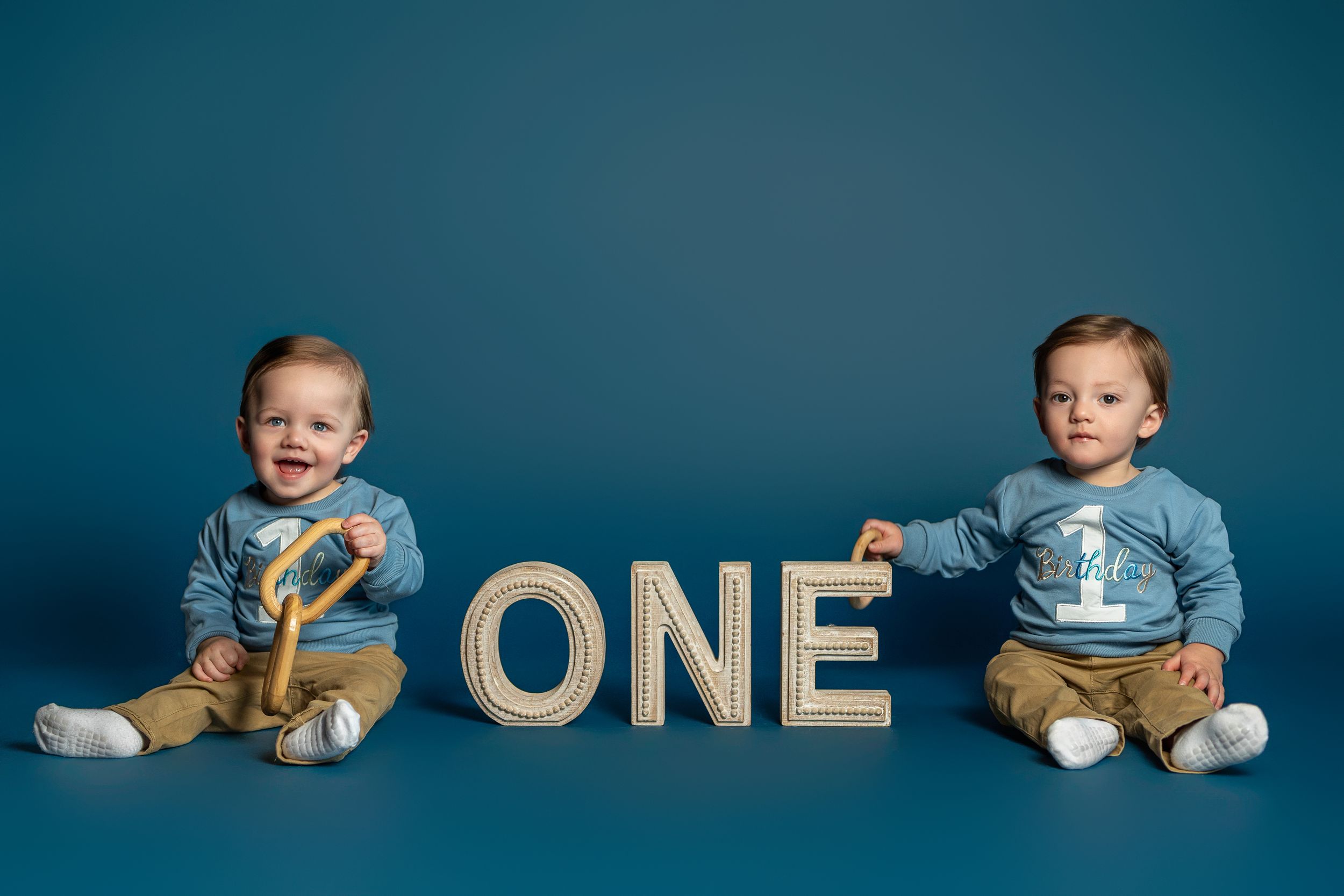 A baby in blue outfit sits next to gold and blue decorative letters and numbers for a first birthday photo session.