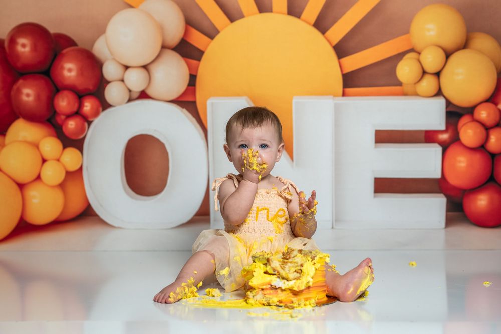 First birthday cake smash setup with sun backdrop and balloon arch in orange and yellow tones.