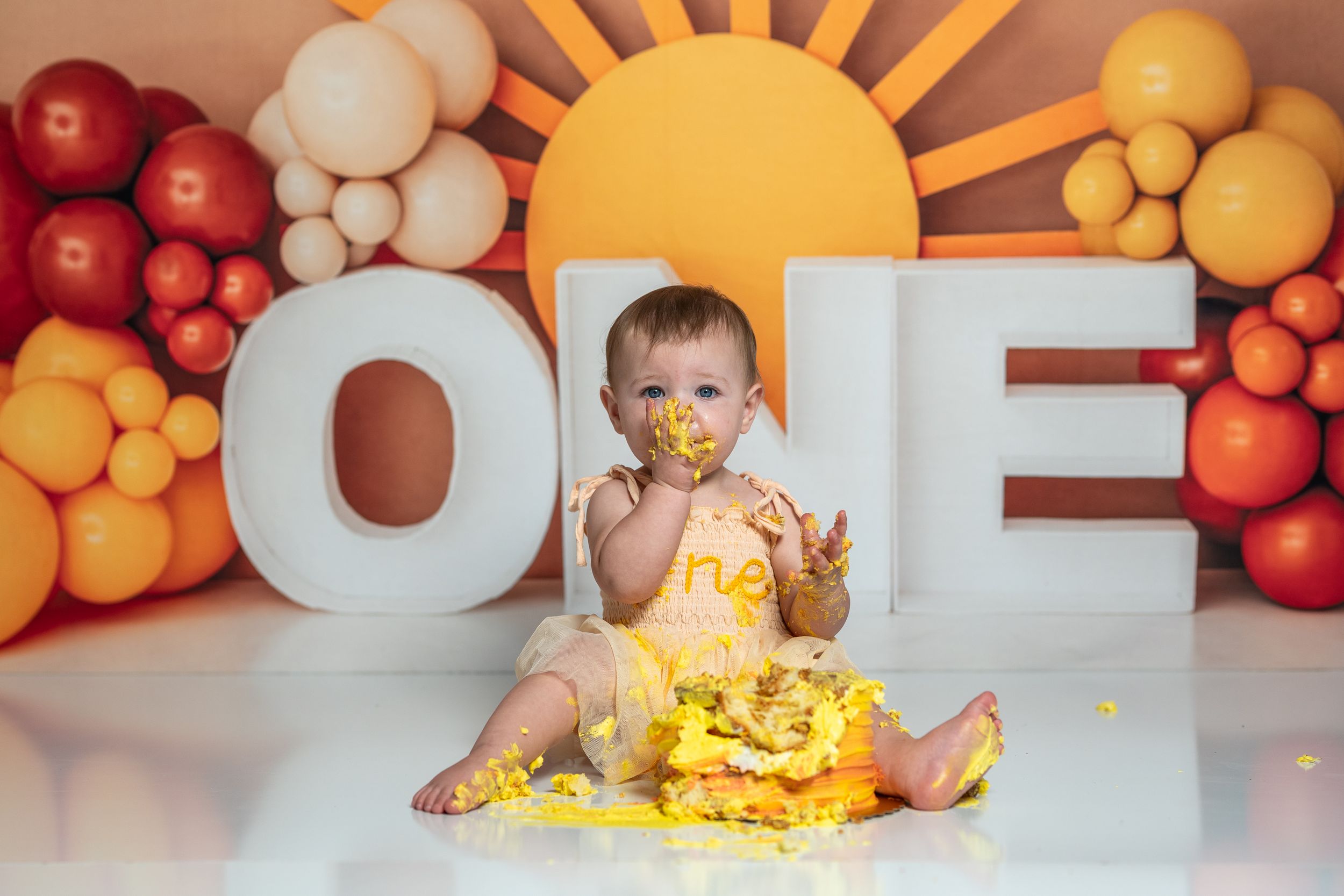 First birthday cake smash setup with sun backdrop and balloon arch in orange and yellow tones.