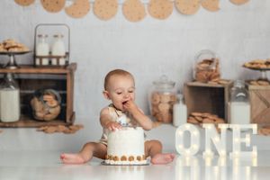 Baby enjoys smashing white birthday cake next to illuminated 'ONE' sign.