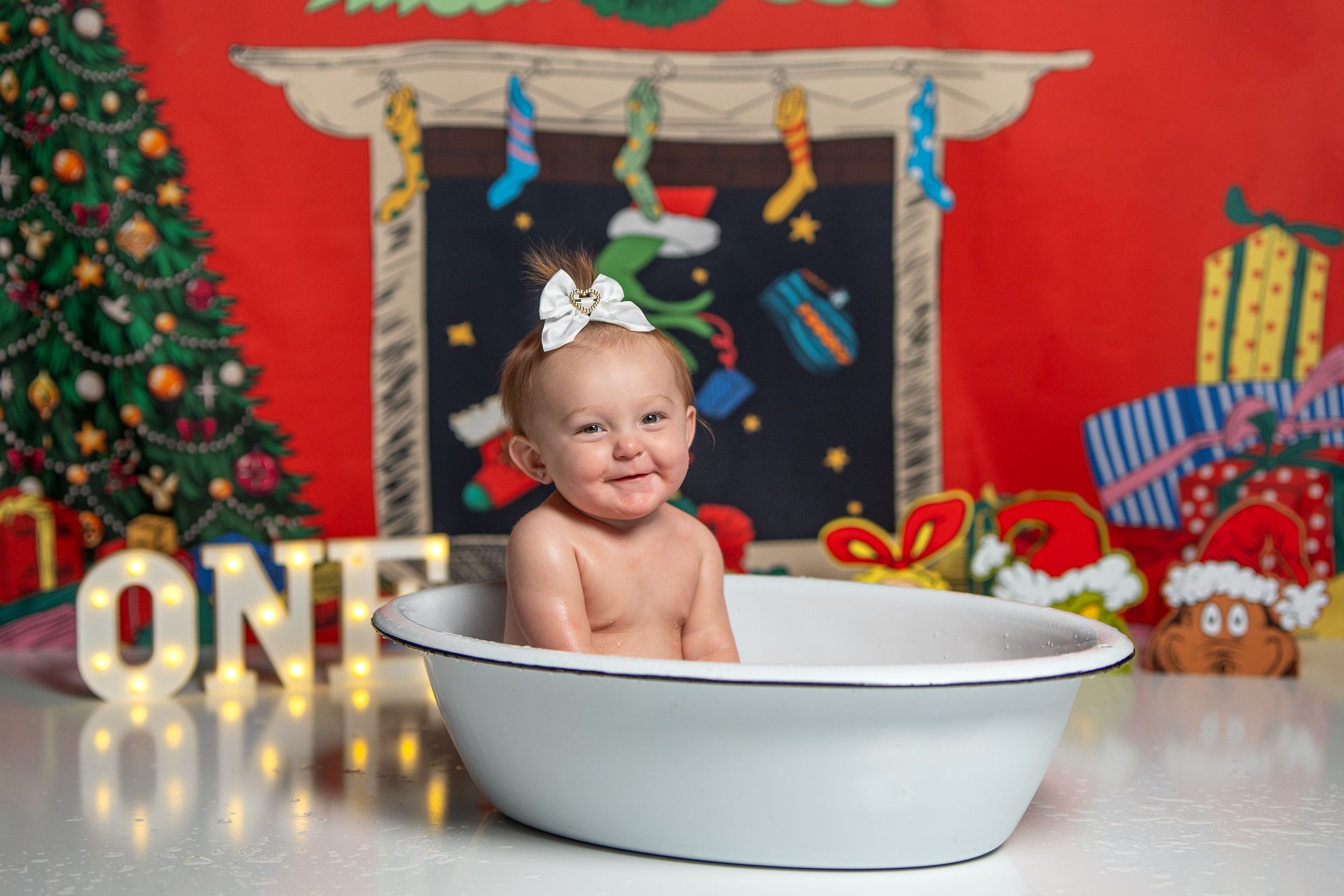 Baby sitting in white bathtub with Christmas decorations and trees in festive red background.