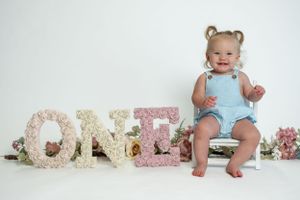 Baby in blue romper sits next to floral 'ONE' letters for first birthday photo.