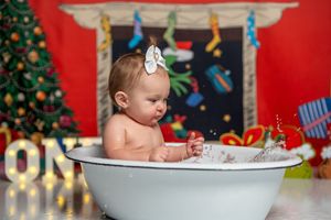A baby sits in a white bathtub during a Christmas-themed photoshoot with decorated trees and red backdrop.