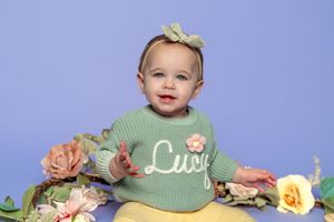 A baby wearing a mint green sweater poses with soft peach and yellow roses against a purple backdrop.