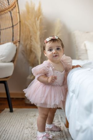 Baby in pink tulle dress standing by bed with boho-styled decor and pampas grass.