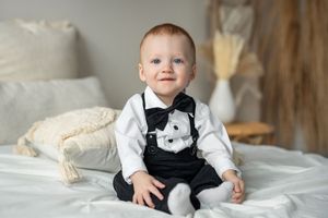 A baby wearing formal attire sits on white bedding while smiling and playing with a stuffed animal.