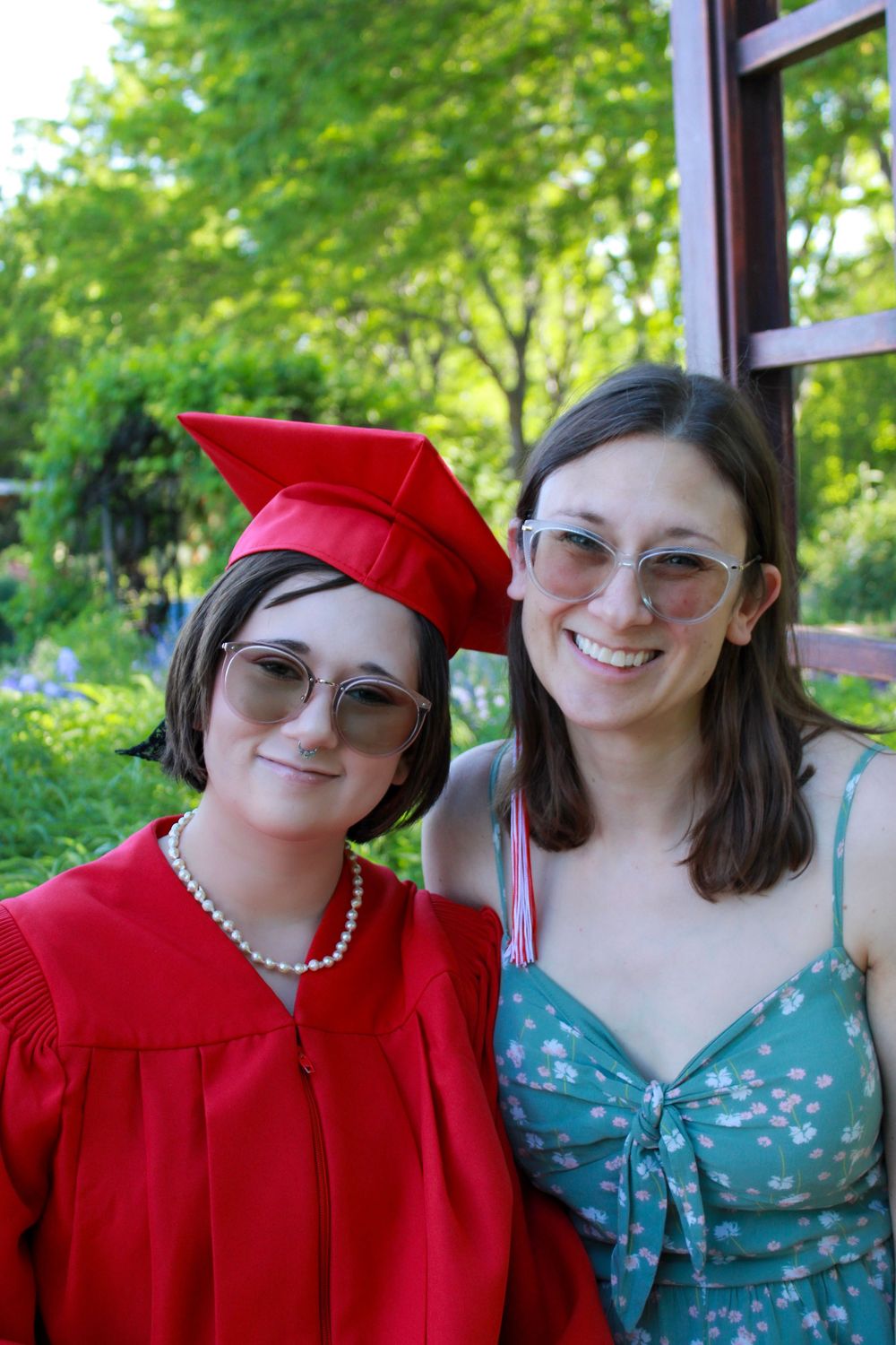 Two people smile together outdoors on graduation day, with one wearing a bright red cap and gown with pearls.
