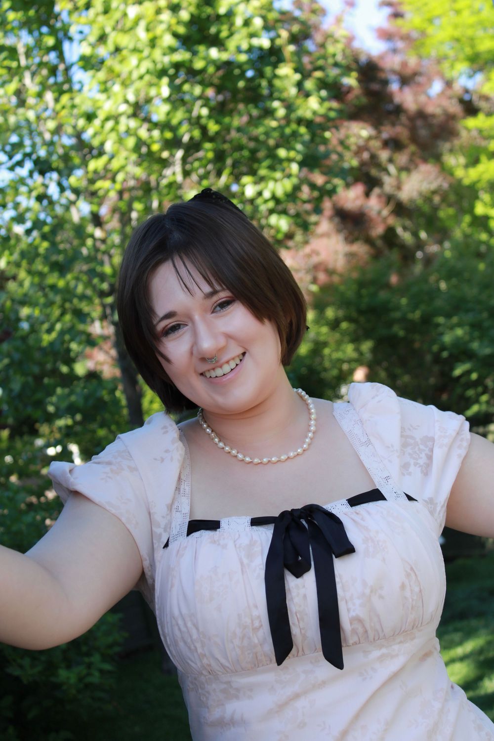 A person in a white dress with a black bow and pearl necklace smiles outdoors in front of green foliage.