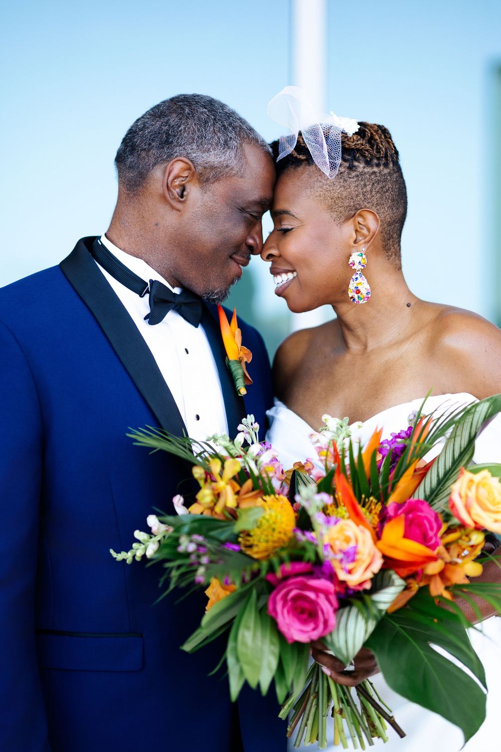 Smiling bride and groom touch foreheads, holding a vibrant tropical bouquet of orange, pink and yellow blooms with greenery.