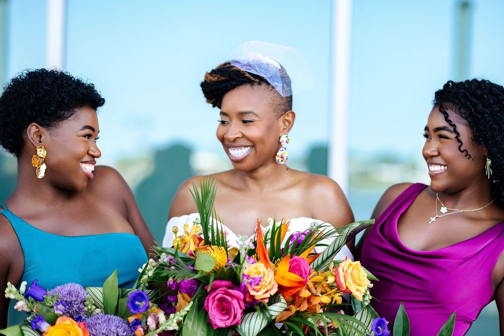 Three smiling women in teal and magenta dresses pose behind a vibrant bouquet of pink, orange, and purple flowers at a wedding celebration.