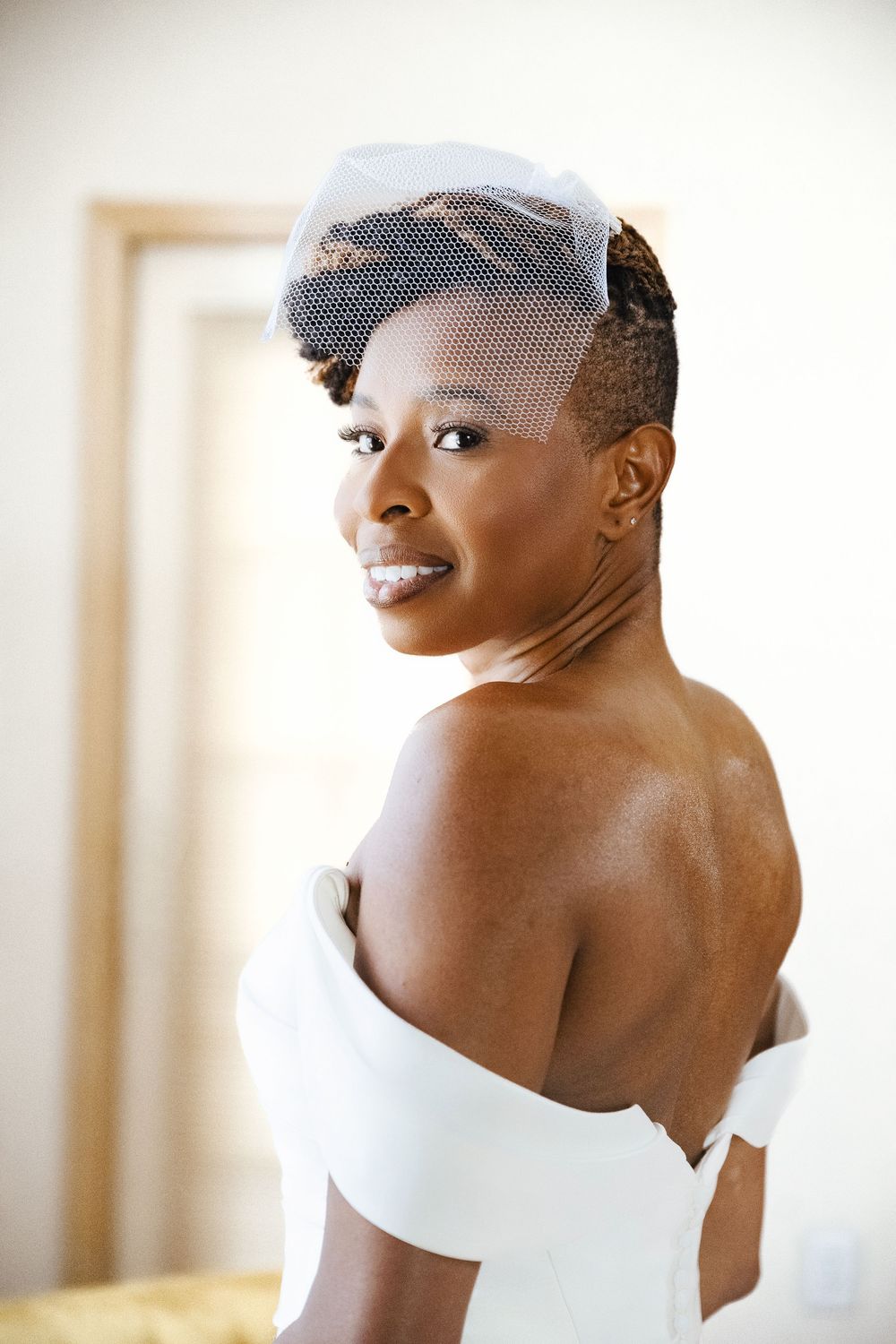 Smiling bride in an off-the-shoulder white wedding gown and birdcage veil glances back over her bare shoulder in a softly lit room.