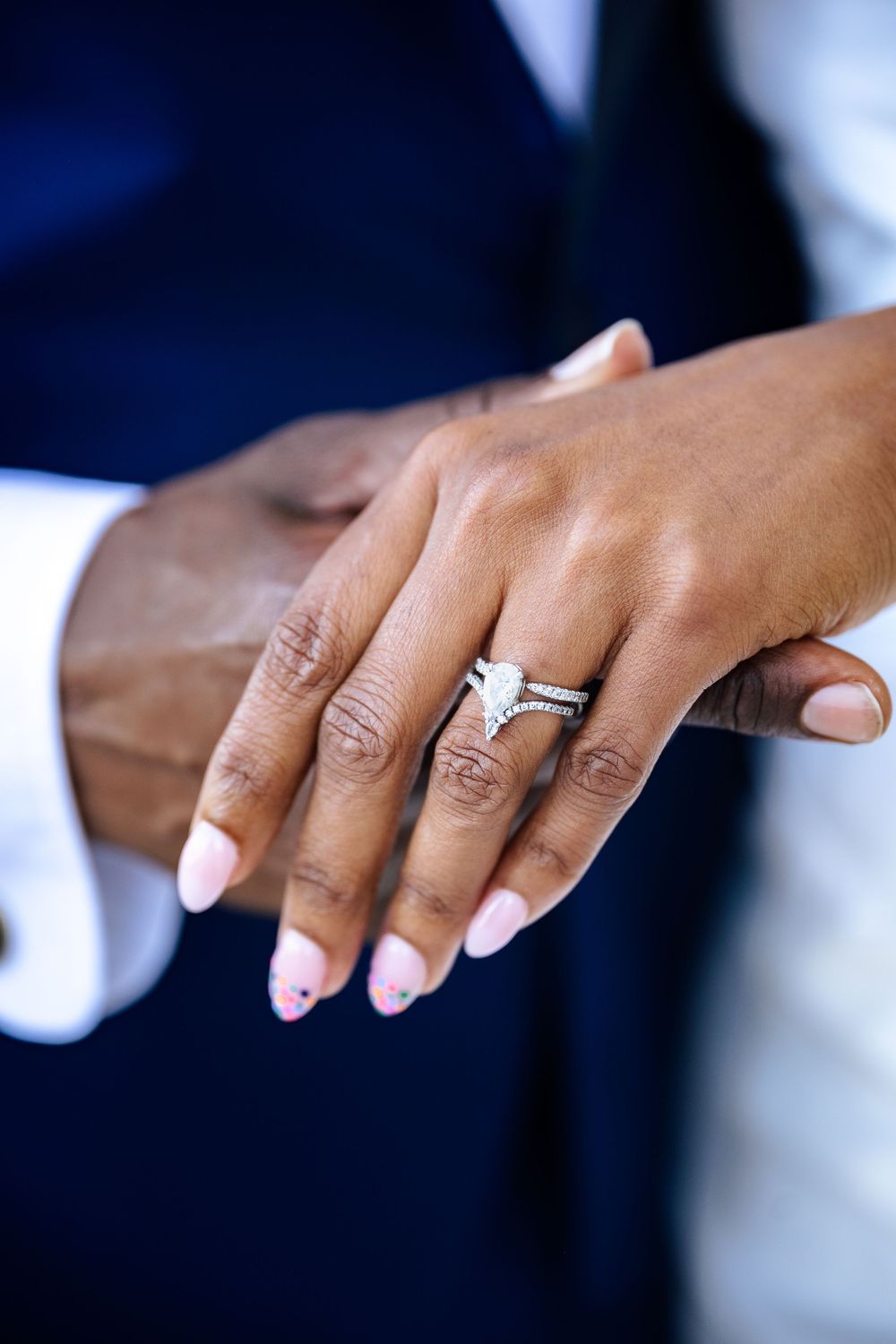 Close-up of a woman's hand with pink manicured nails and a pear-shaped diamond engagement ring resting on a man's hand in a navy suit.
