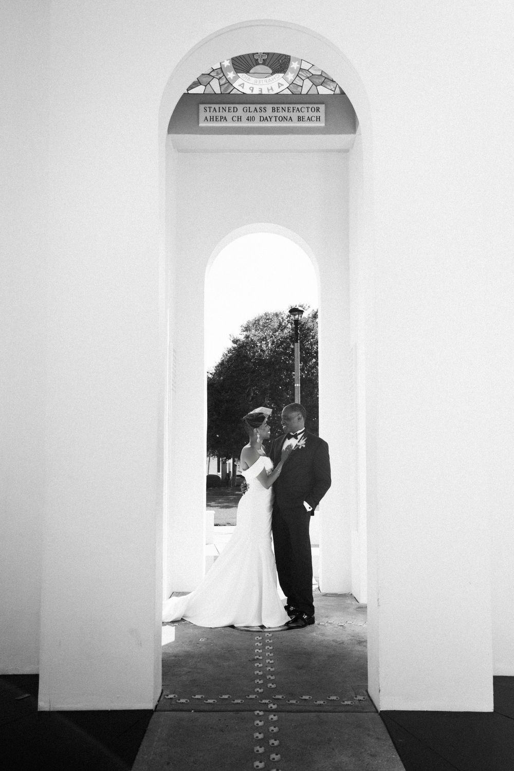 Bride in a flowing white gown and groom in a dark suit embrace beneath a white arched passageway with stained glass in black and white.