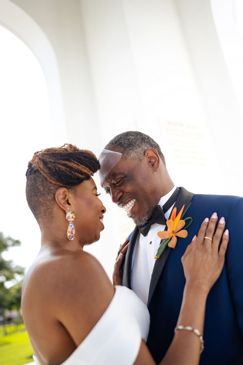 Smiling bride in an off-shoulder white gown and groom in a navy tuxedo with an orange floral boutonniere share a joyful moment outdoors.