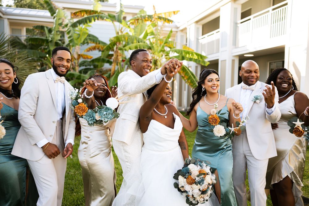Joyful wedding celebration with bridal party wearing white and sage green attire posing outdoors near tropical plants.