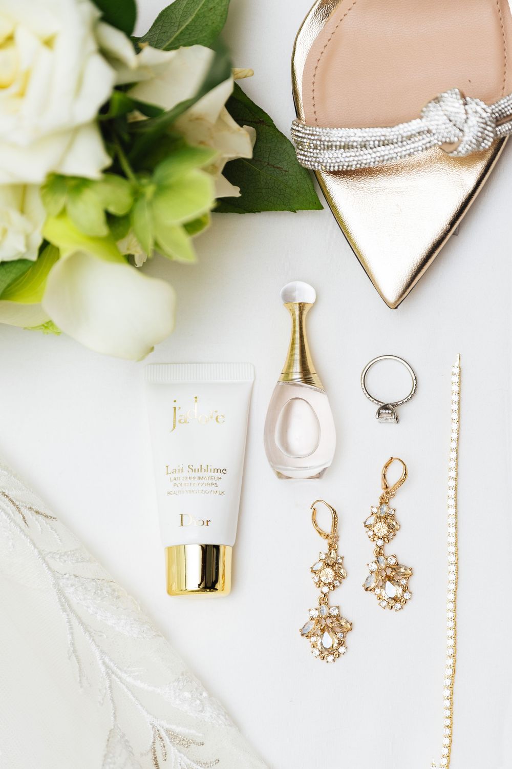 Elegant bridal flat lay with Dior J'adore perfume, gold earrings, ring, heels, and white flowers on white background.