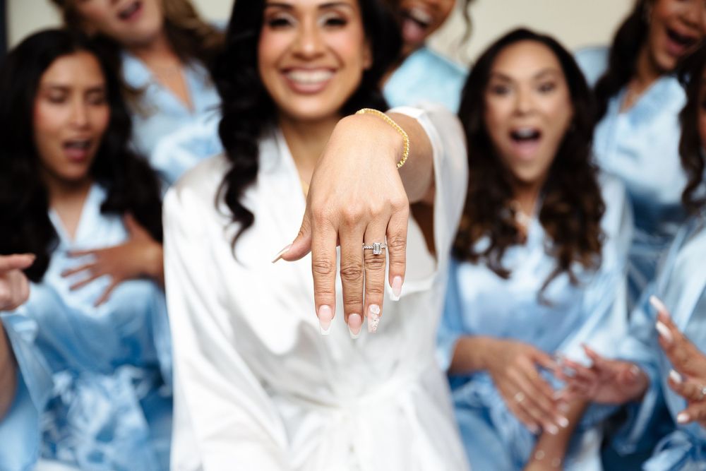 Bride shows off engagement ring surrounded by excited bridesmaids in light blue robes during bridal party preparations.