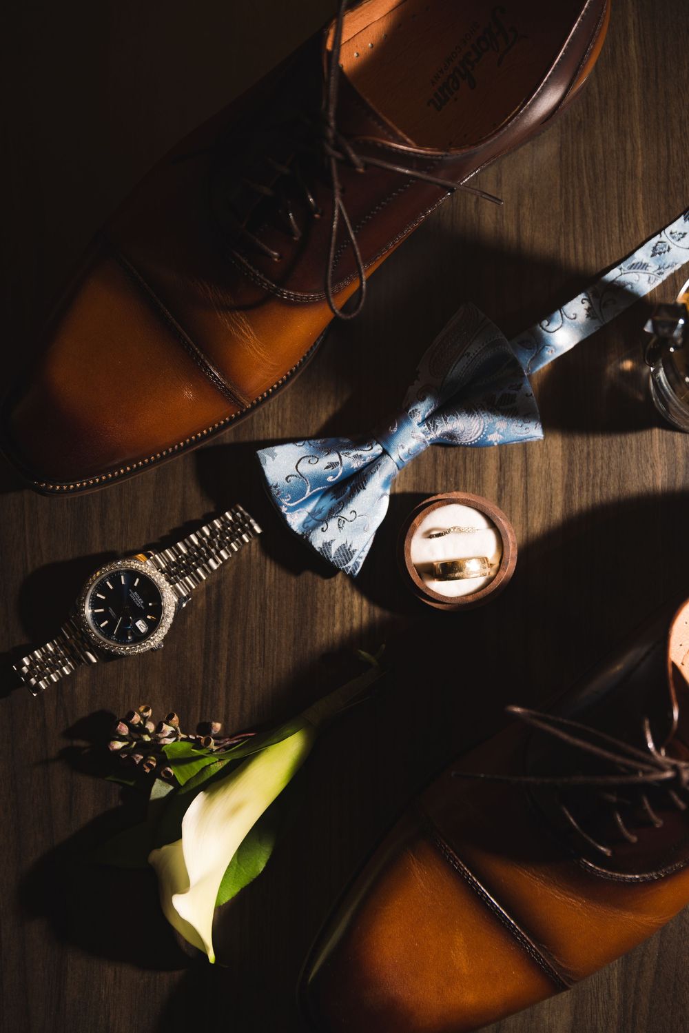 Groom wedding accessories flat lay with brown dress shoes, blue bow tie, watch, boutonniere, and gold ring on wood floor.