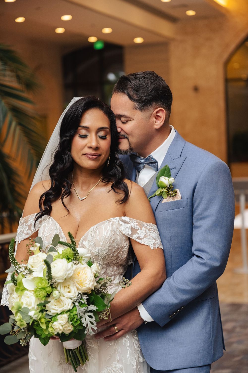 A bride and groom share a tender moment, she holds white and green bouquet, he wears a blue suit with boutonniere.