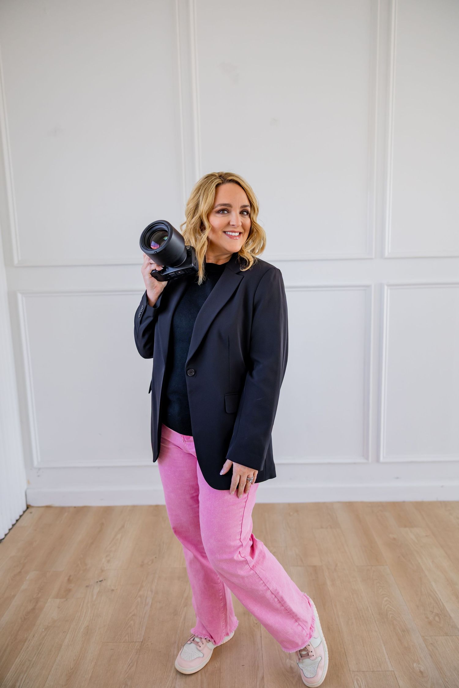 Woman in black blazer and pink pants poses playfully with camera in studio setting.