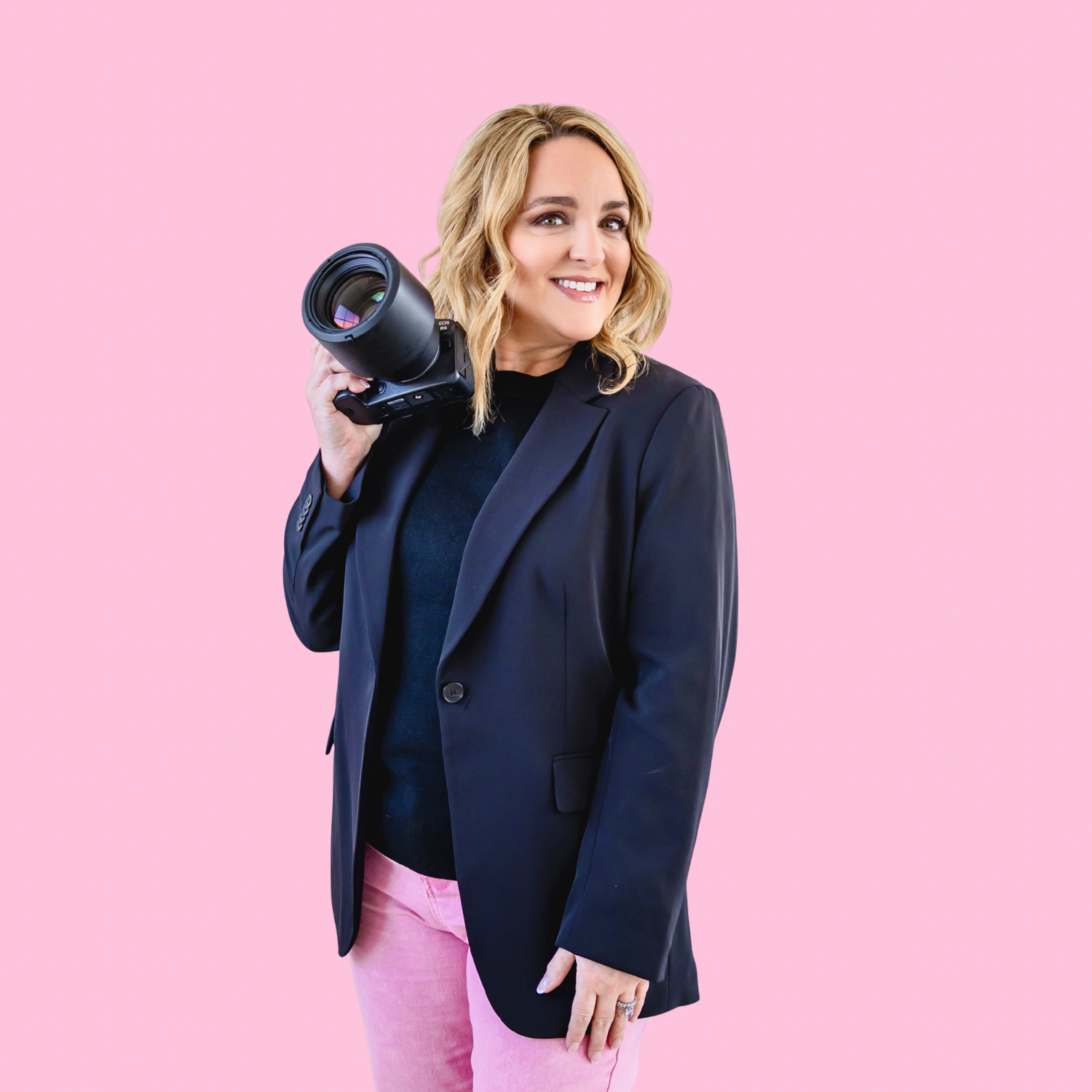 Woman in black blazer and pink pants poses playfully with camera in studio setting.