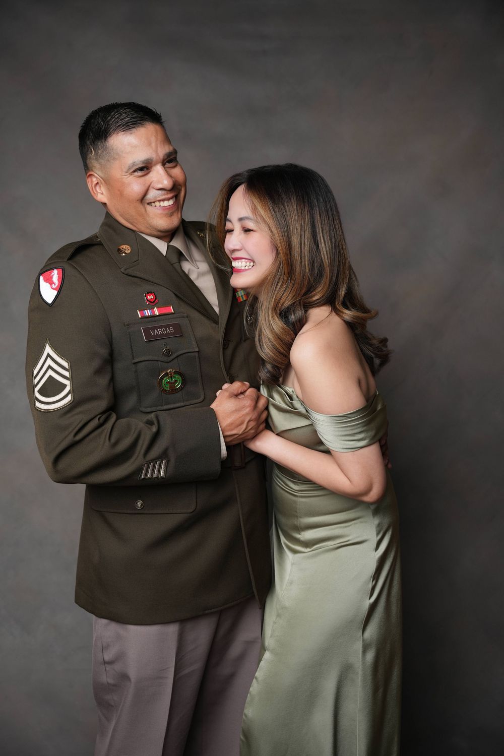 Hispanic couple embracing and laughing in formal wear in front of a brown backdrop