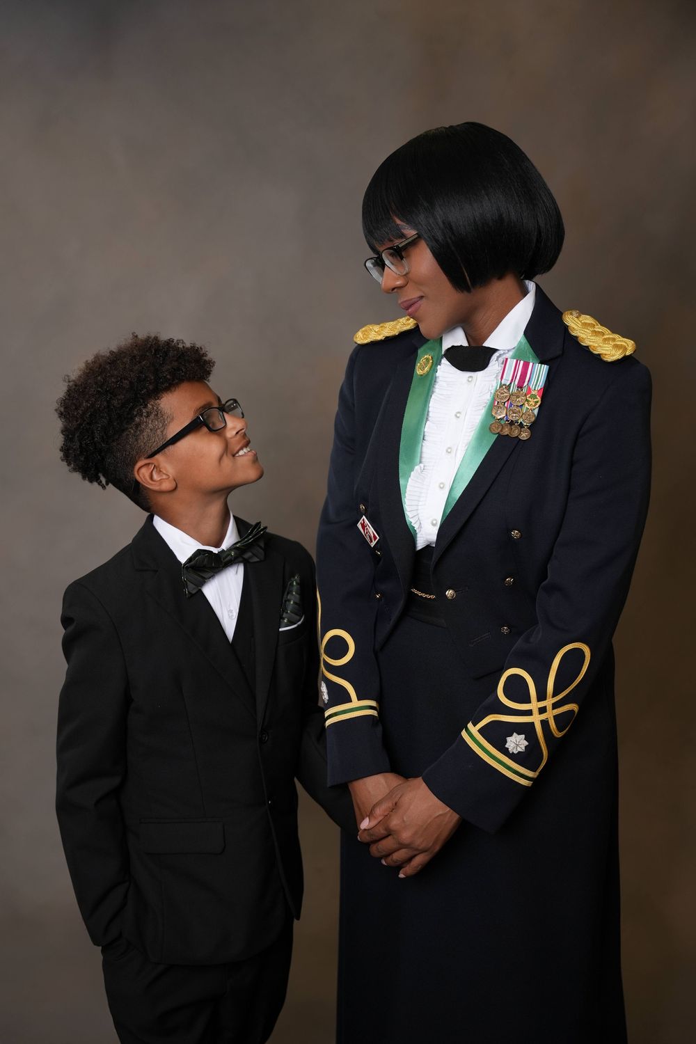 picture of a black mother and son in military uniform and son in a suit and bow tie posing in front of a brown photography backdrop.