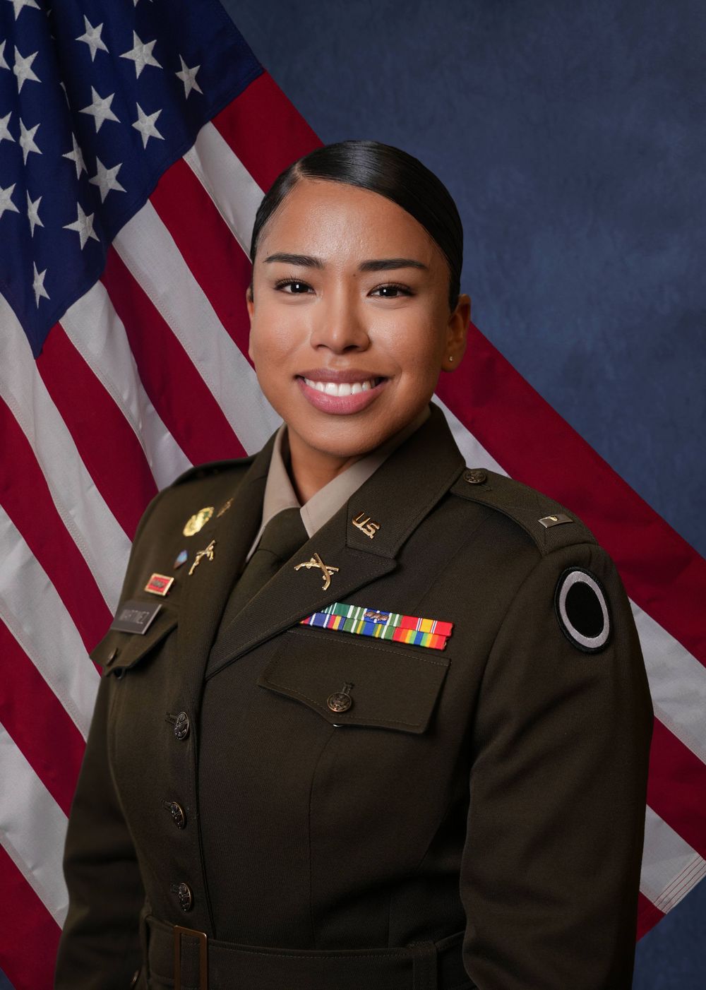 Picture of a hispanic woman in Army dress uniform posing in front of a blue backdrop with American flag draped behind.