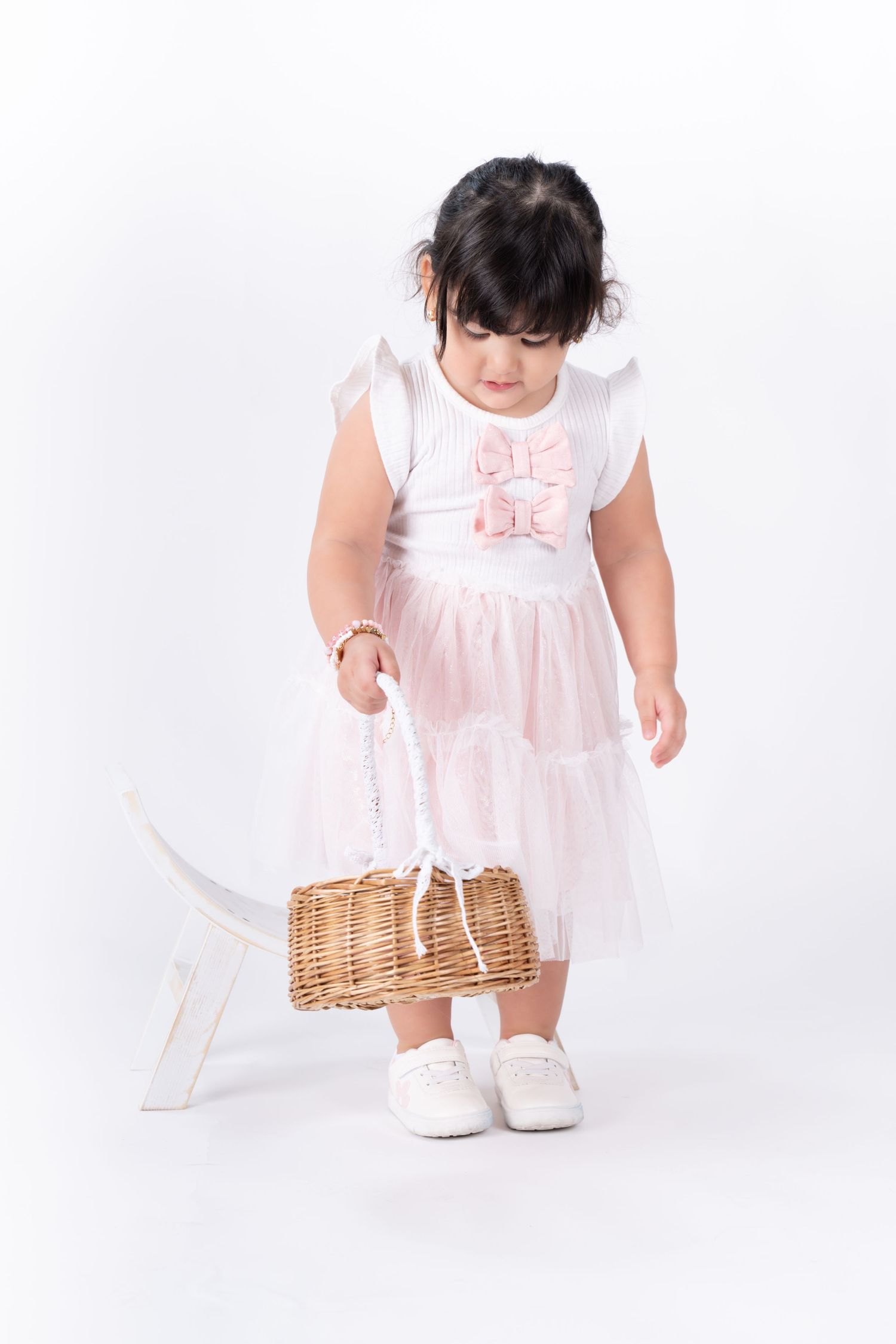 Small child in white dress with pink bows holds wicker basket.