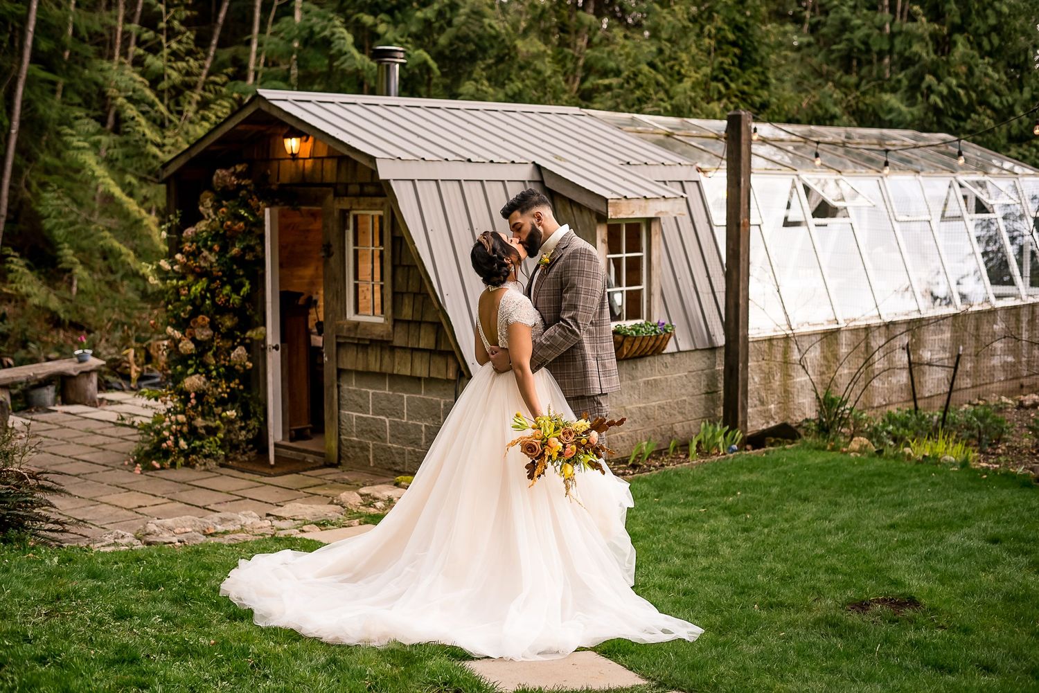 A bride and groom kiss on the hillside in front of their greenhouse wedding venue. The bride holds her bouquet of autumn coloured flowers.