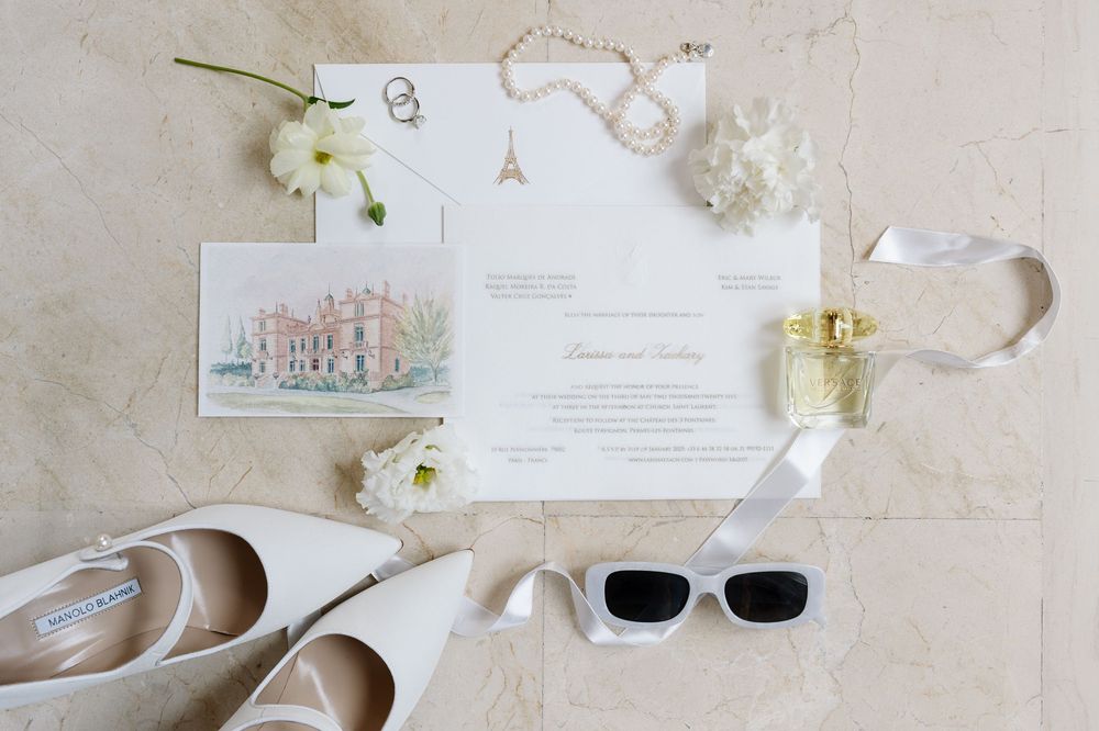 Wedding invitation flatlay with white shoes, sunglasses, and floral accents on marble background.