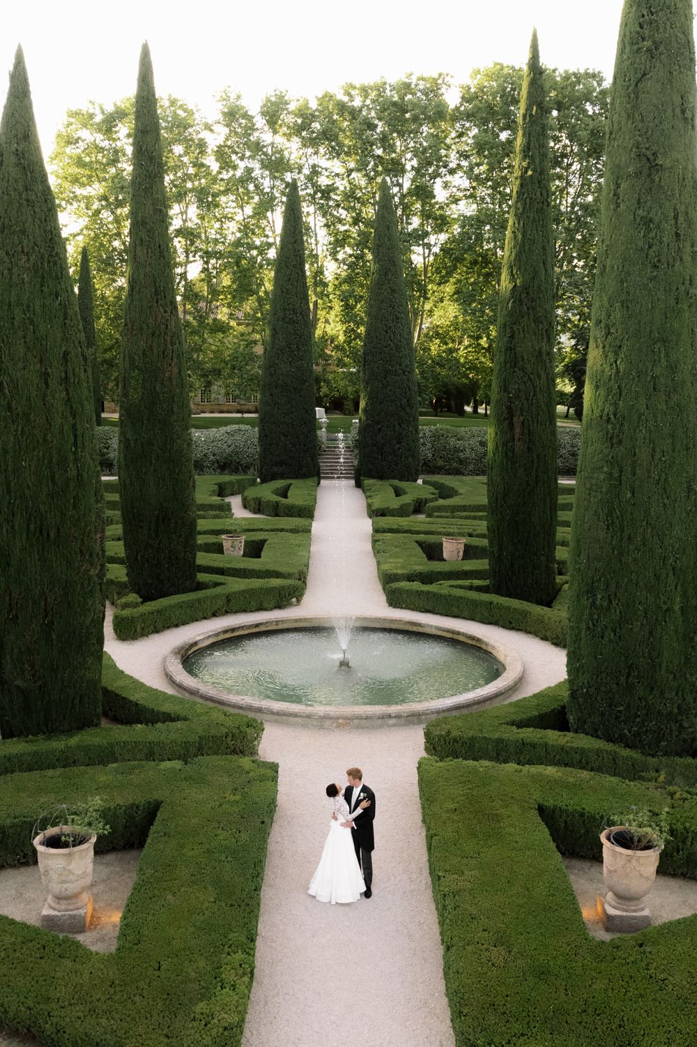 Aerial view of newlyweds in formal garden with cypress trees and fountain.