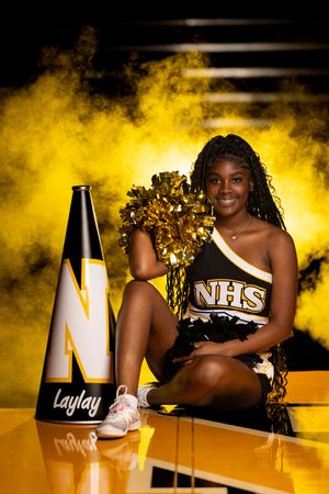 A cheerleader in NHS uniform poses with gold pom poms and megaphone against dramatic yellow smoke background.
