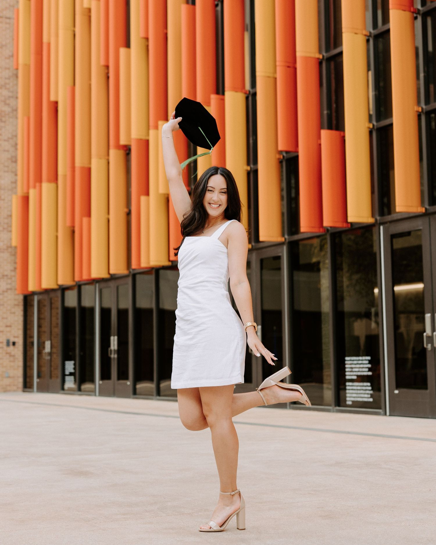 San Antonio Graduation Photography. Woman in white dress poses playfully against modern orange architectural backdrop.