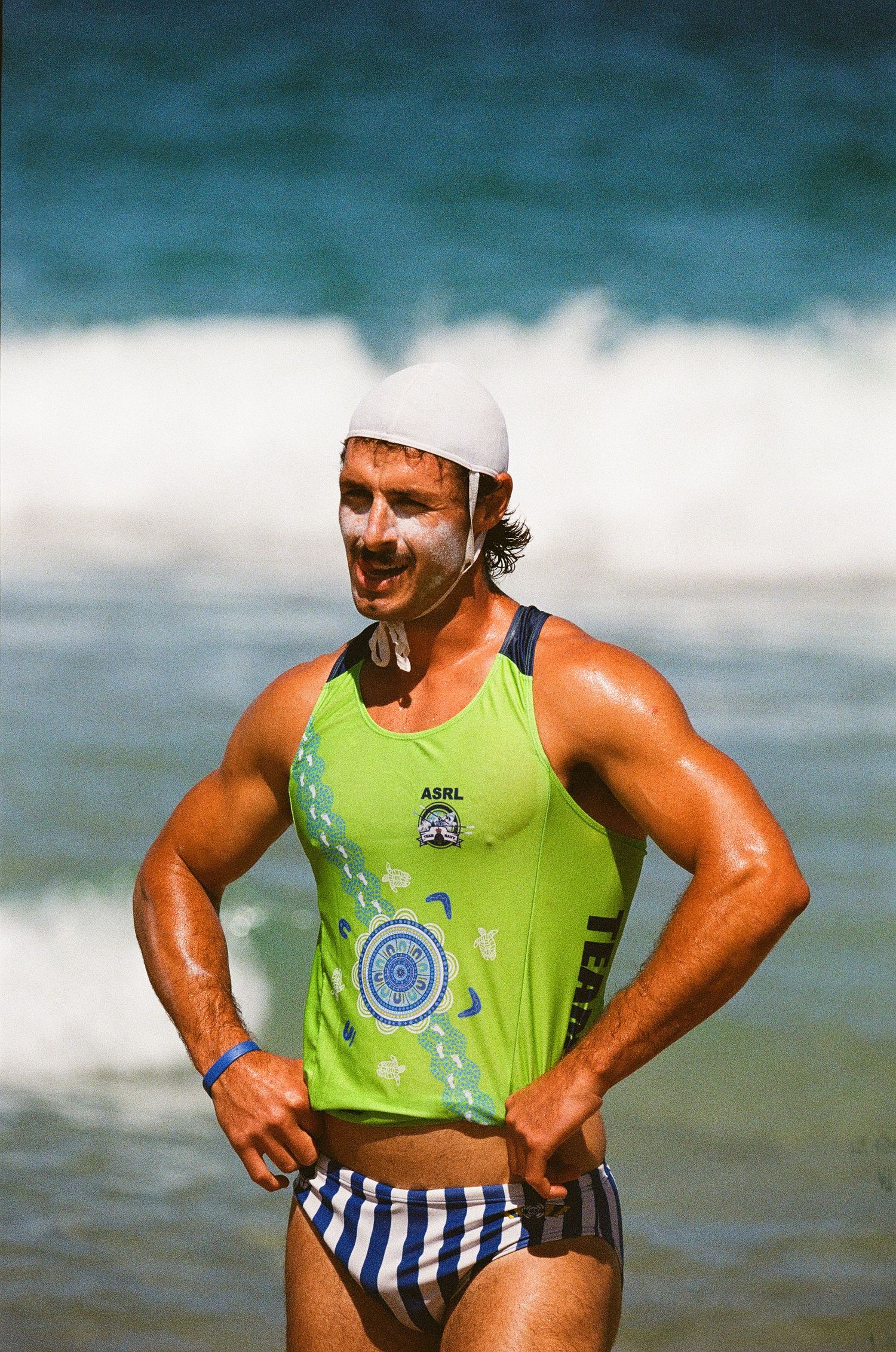 Athletic lifeguard in bright green tank top and striped swimwear poses on sunny beach with ocean waves behind.
