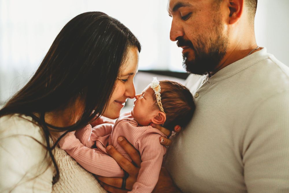 Parents share an intimate moment with their baby wearing pink clothing in a warmly lit indoor setting.