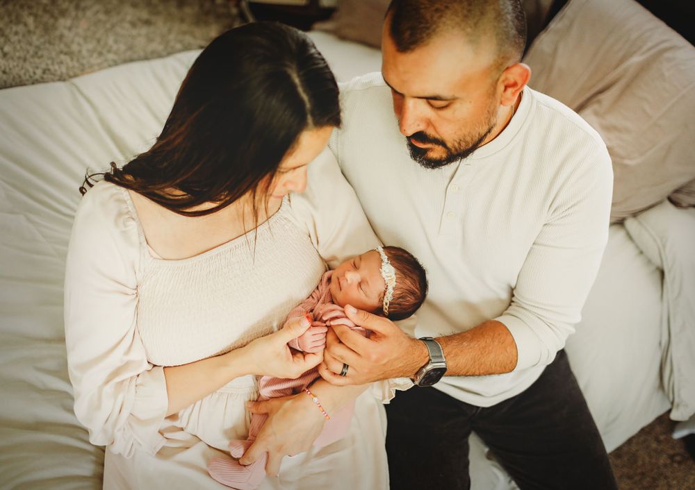 Two new parents in white sweaters lovingly gaze at their newborn baby while sitting together on a bed.
