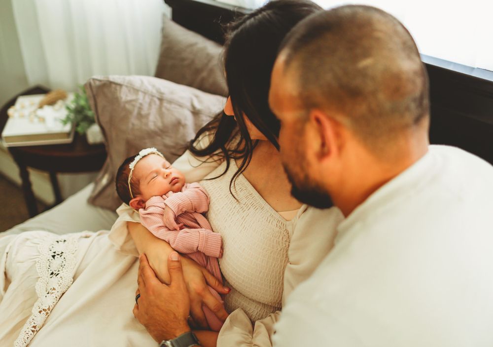 Two parents lovingly hold and gaze at their newborn baby while sitting together on a bed.