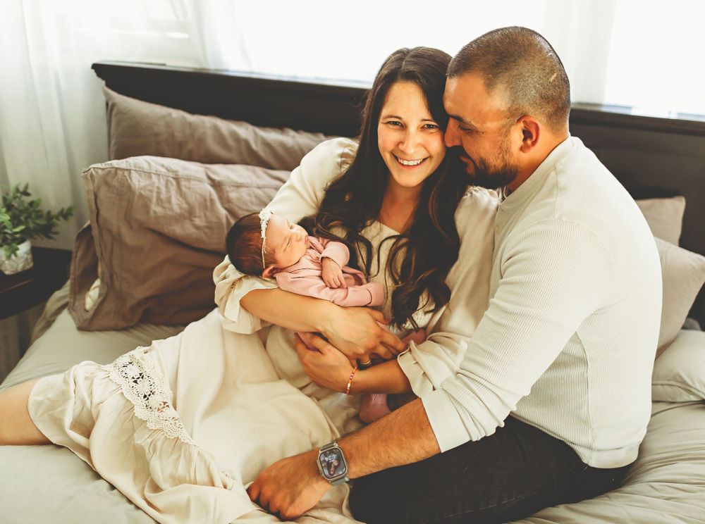 A loving couple sits on a bed tenderly cradling their newborn baby in a cozy bedroom setting.