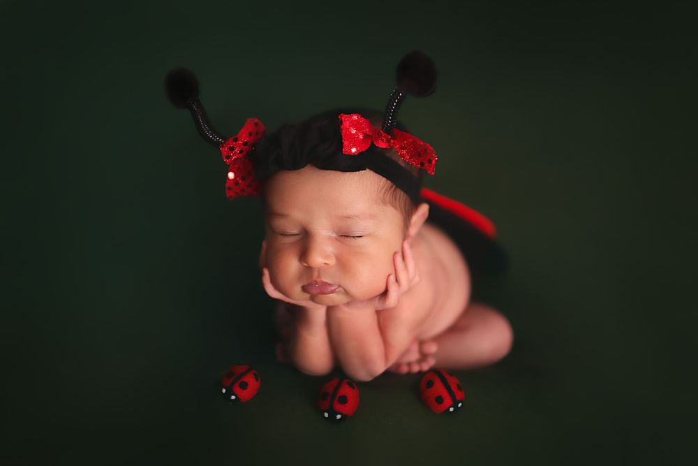 Newborn wrapped in red ladybug costume with black spots and antennae rests peacefully on dark green backdrop.