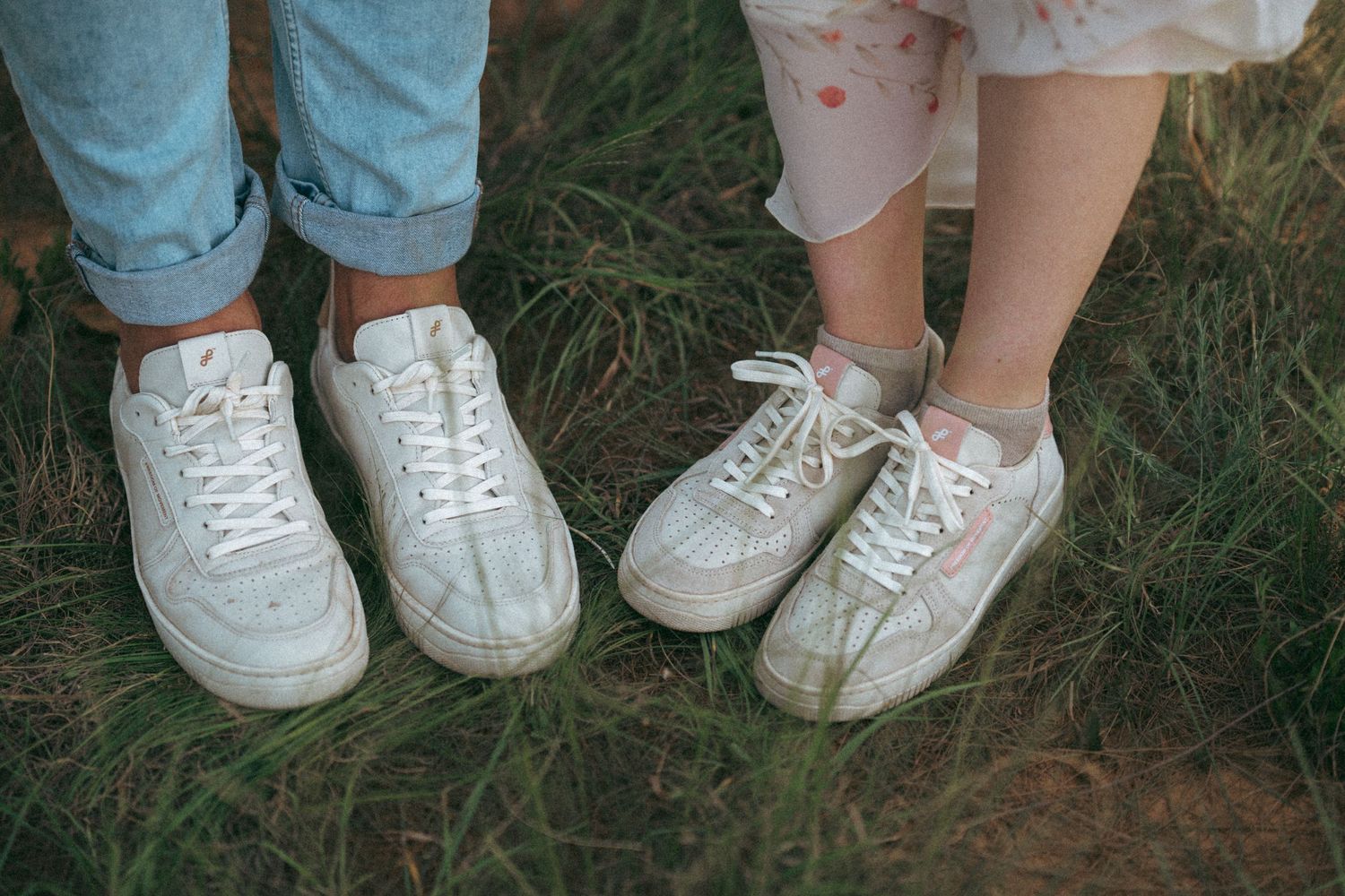 Two pairs of matching white sneakers photographed side by side on grass.
