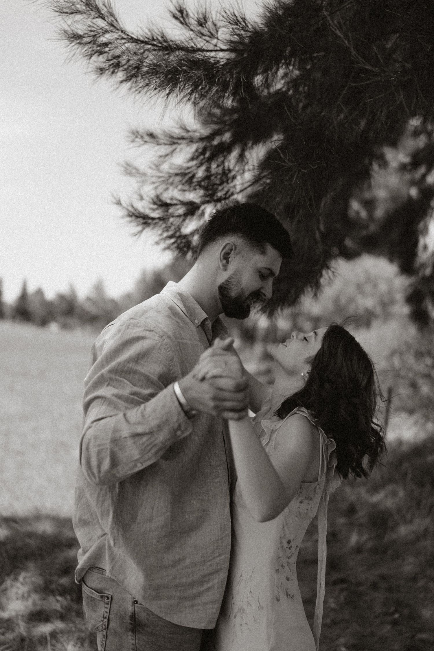 A romantic black and white portrait of a couple dancing under a pine tree.